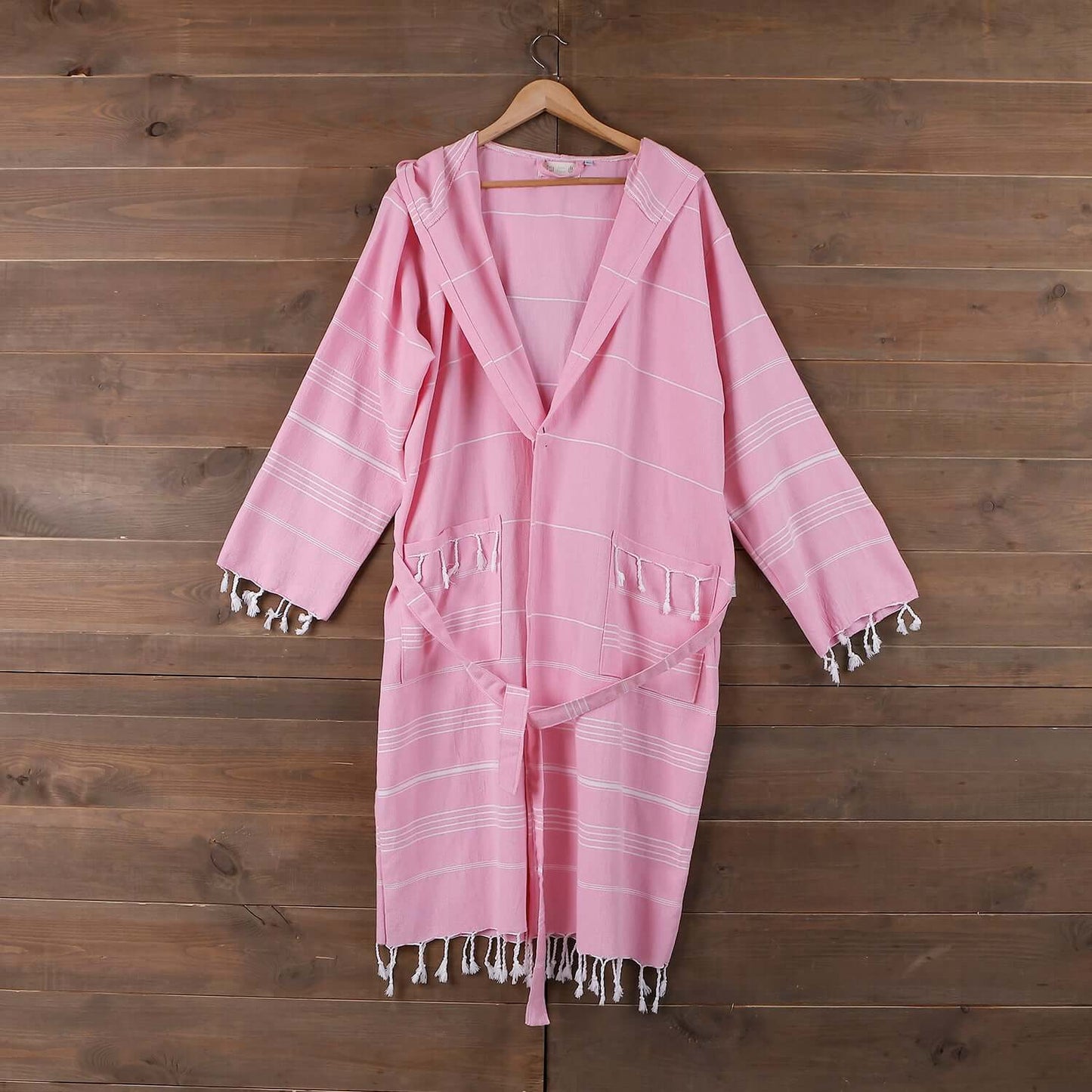 The Loom Legacy medium/large pink bathrobe hangs gracefully on a wooden hanger against a rustic wooden backdrop. The bathrobe boasts a calming blue shade with white stripe patterns, a tie belt for a secure fit, and fringe detailing along the pockets and