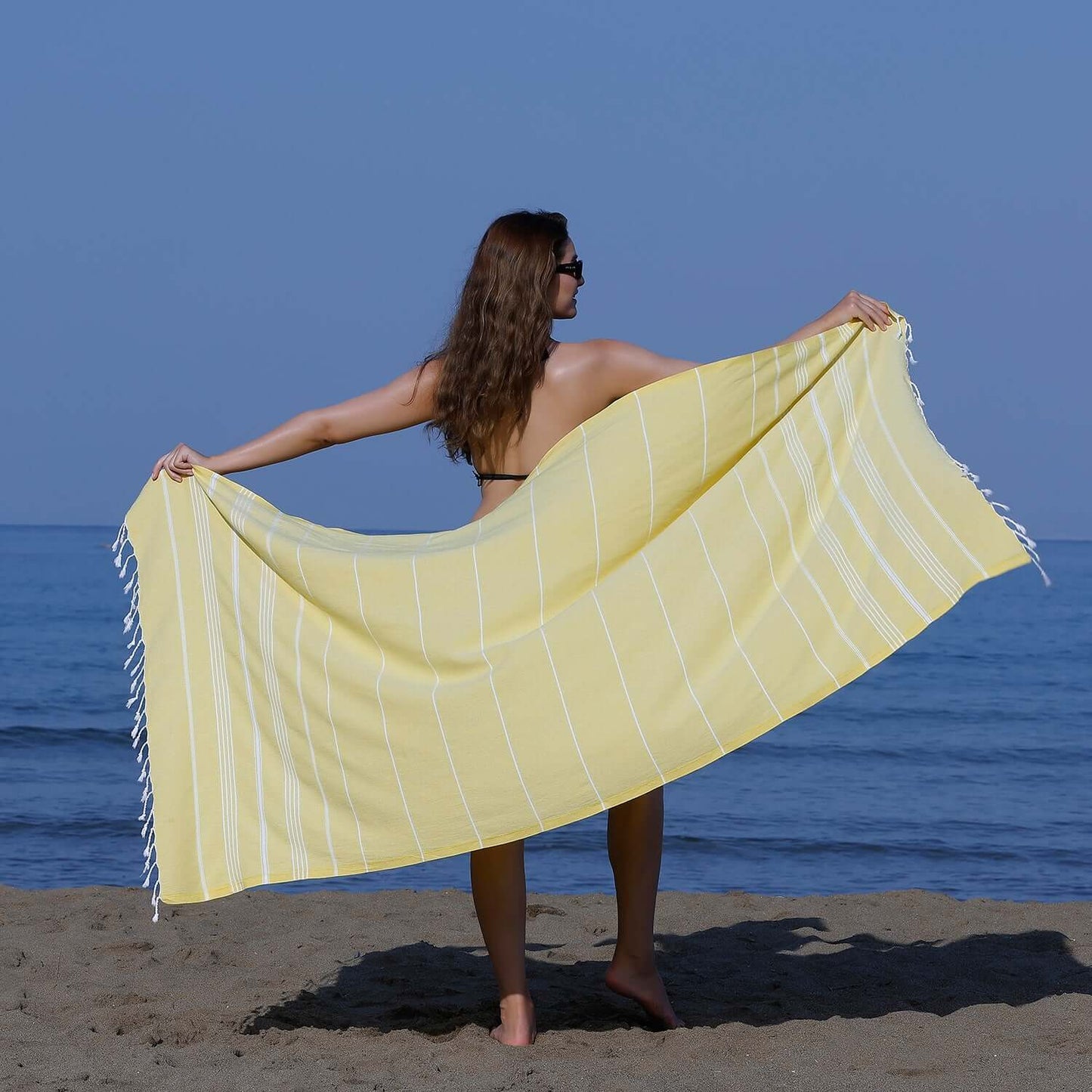 Woman at the beach showcasing a Loom Legacy yellow striped beach towel with tassels, with the sea in the background