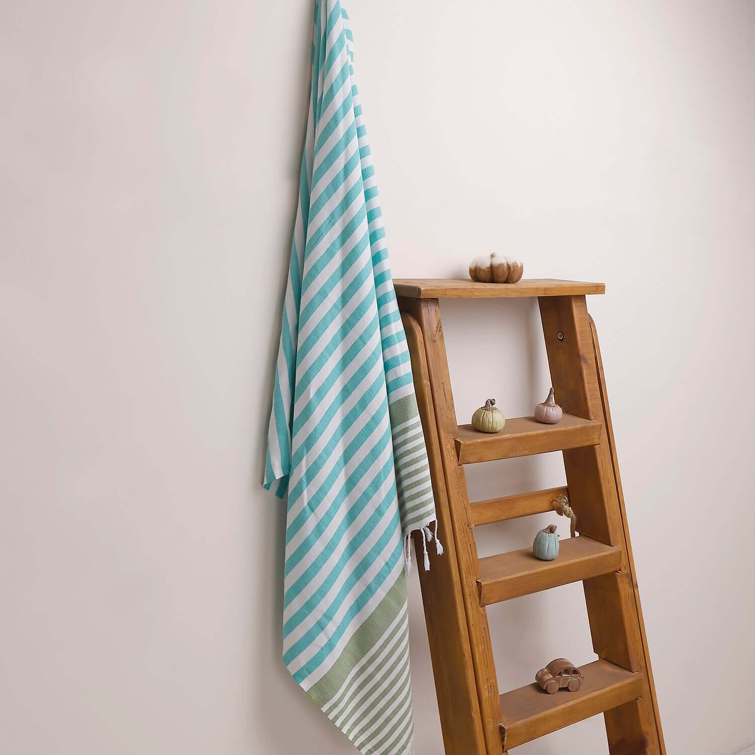 Loom Legacy turquoise and green striped beach towel with tassels, draped over a wooden ladder beside small decorative items on the shelves.