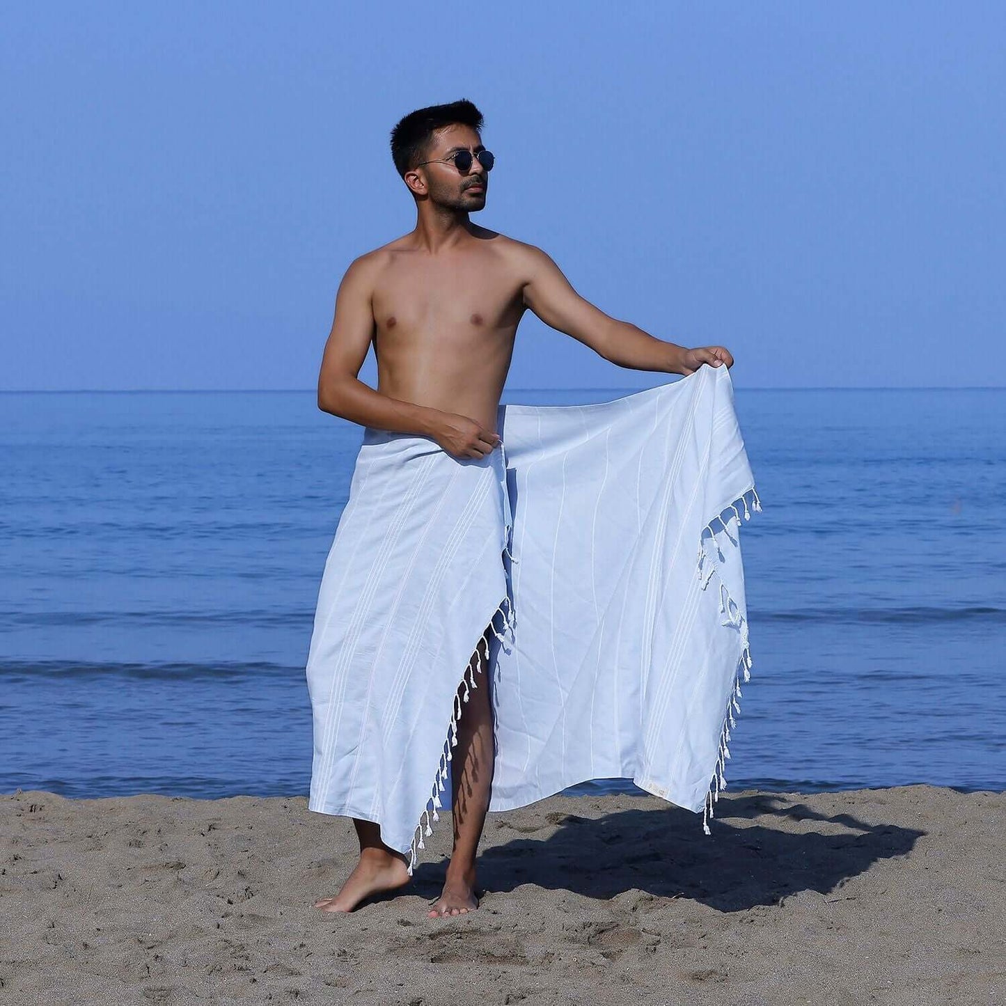 Man at the beach showcasing a Loom Legacy light blue striped beach towel with tassels, with the sea in the background.