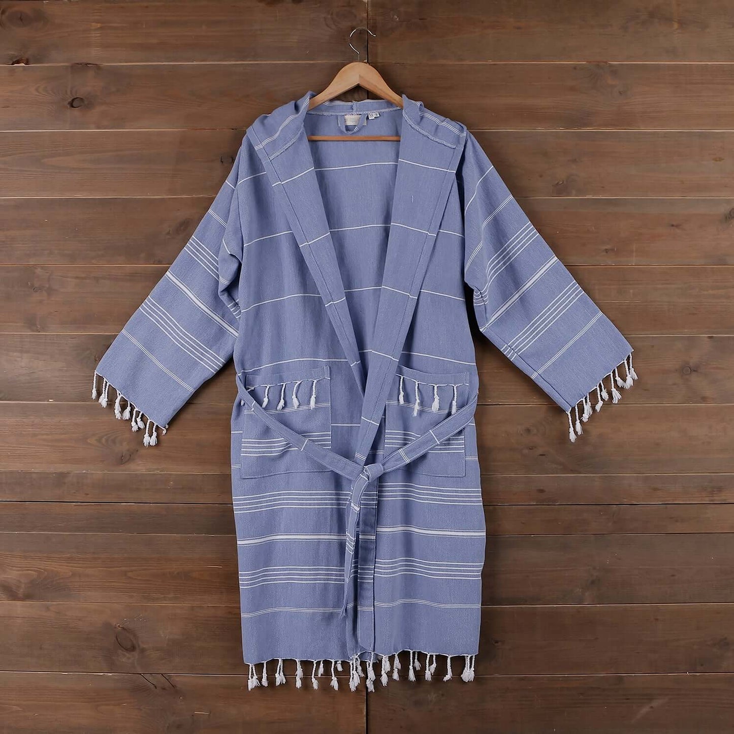 The Loom Legacy xsmall/small blue bathrobe hangs gracefully on a wooden hanger against a rustic wooden backdrop. The bathrobe boasts a calming blue shade with white stripe patterns, a tie belt for a secure fit, and fringe detailing along the pockets and h