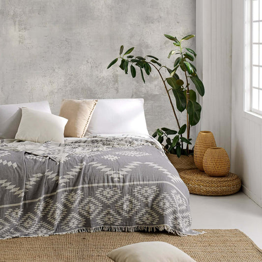 Cozy bedroom interior featuring a Loom Legacy patterned grey cotton blanket with intricate white designs, laid on a comfortable bed, complemented by neutral-toned pillows, a tall potted plant, and woven floor accessories against a rustic gray wall backdro