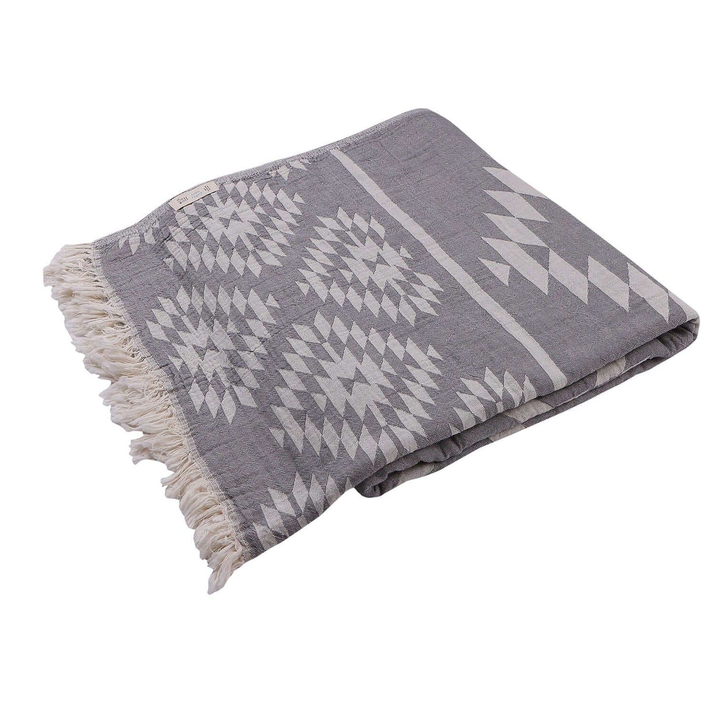 A folded Loom Legacy cotton blanket showcasing a detailed white geometric pattern against a soft grey background, finished with fringe edges, and subtly featuring the brand label