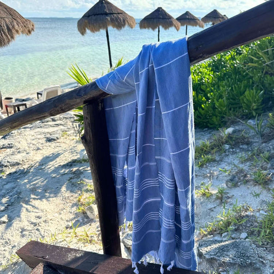 Loom Legacy dark blue and white patterned beach towel over a wooden rail, with a beach background showing straw umbrellas, clear water, and sandy shore.