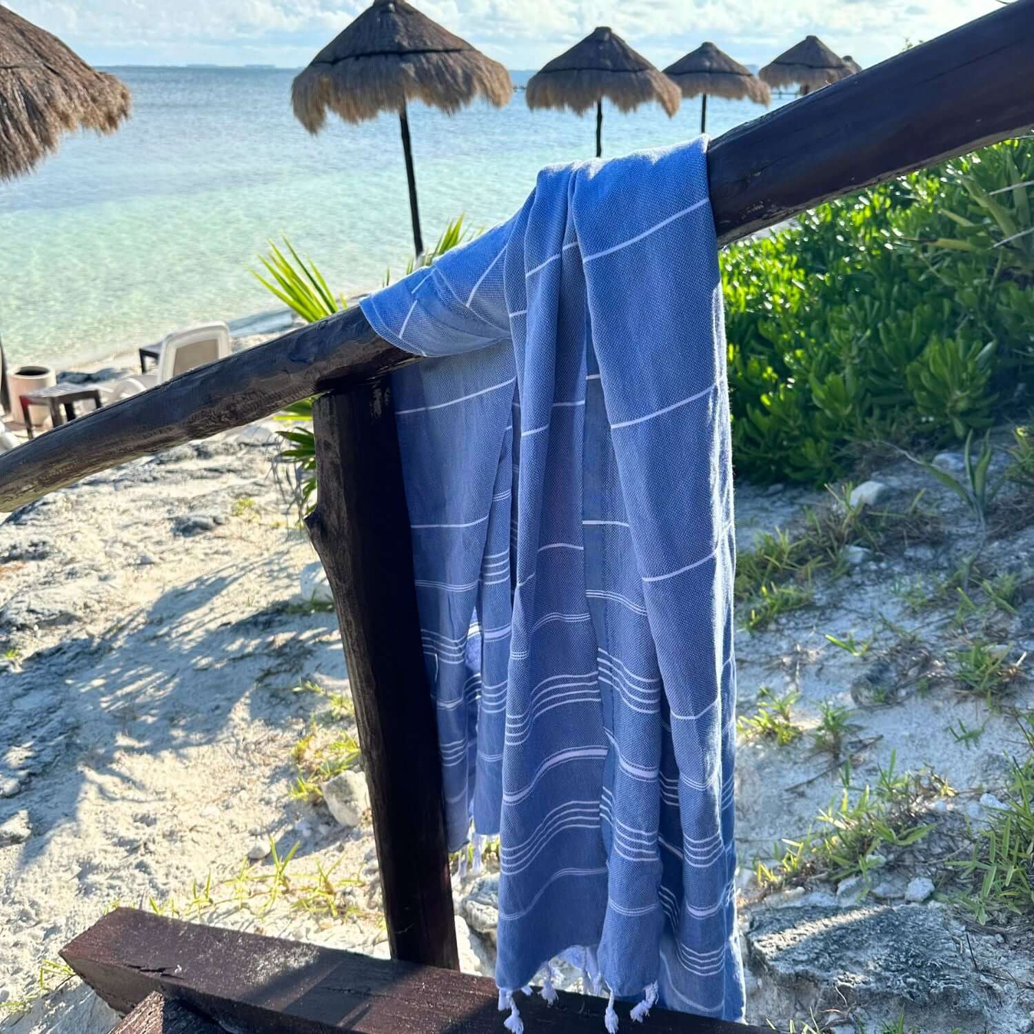 Loom Legacy dark blue and white patterned beach towel over a wooden rail, with a beach background showing straw umbrellas, clear water, and sandy shore.