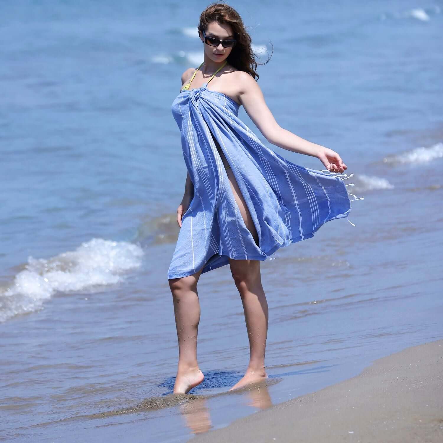 A woman wearing sunglasses stands by the shoreline, draped in a dark blue Loom Legacy beach towel with white stripes, as waves gently lap at her feet
