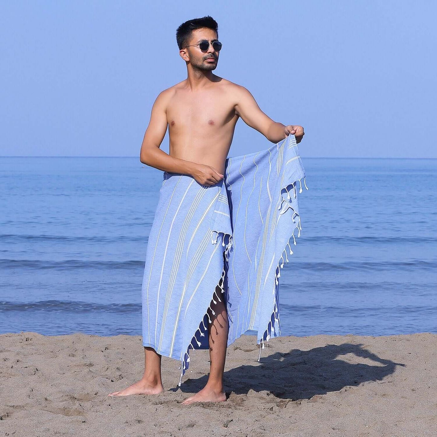 Man at the beach showcasing a Loom Legacy dark blue striped beach towel with tassels, with the sea in the background.
