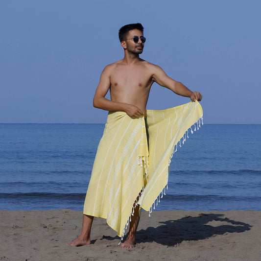 Man at the beach showcasing a Loom Legacy yellow striped beach towel with tassels, with the sea in the background.