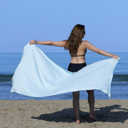 Woman at the beach showcasing a Loom Legacy aqua striped beach towel with tassels, with the sea in the background.