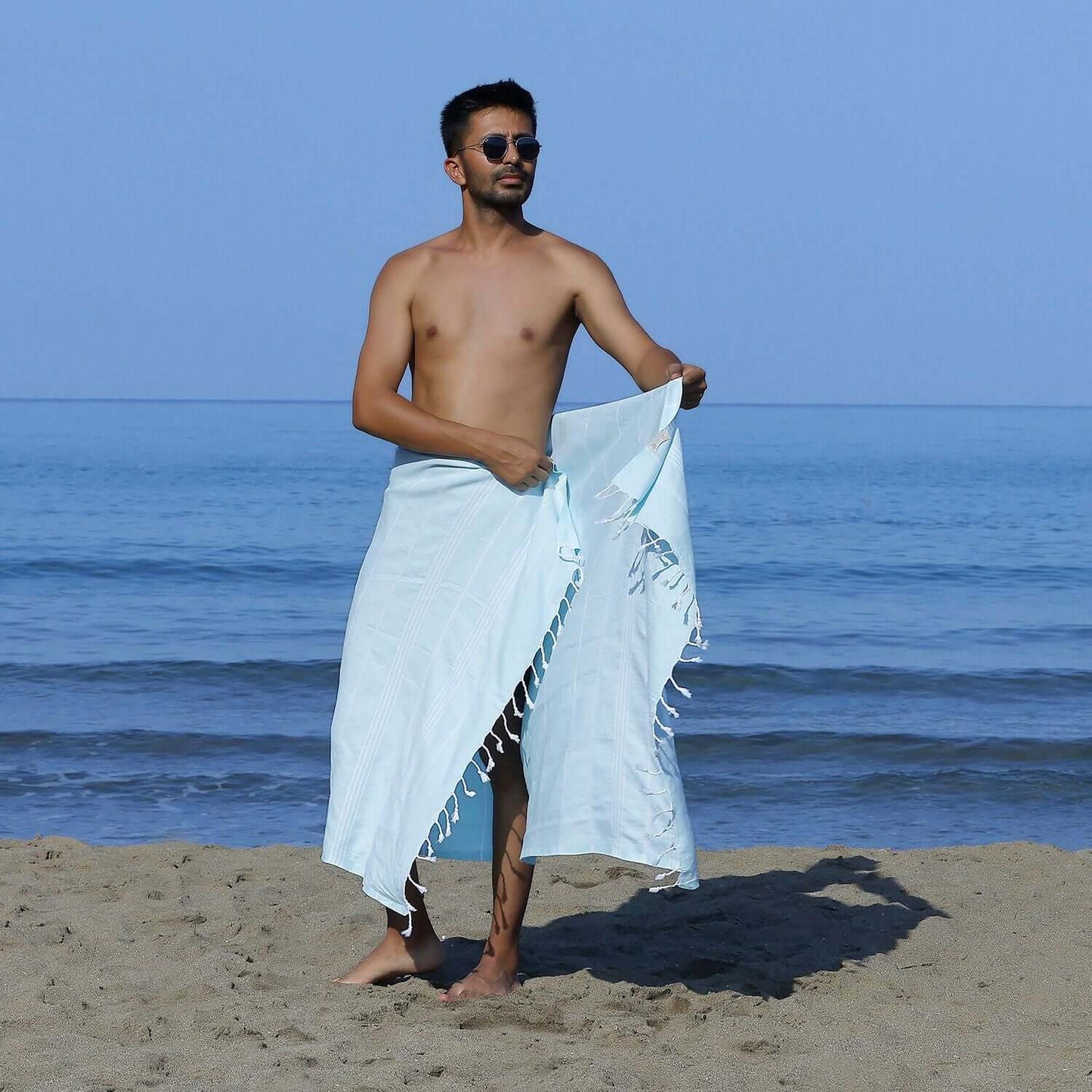 Man at the beach showcasing a Loom Legacy aqua striped beach towel with tassels, with the sea in the background.