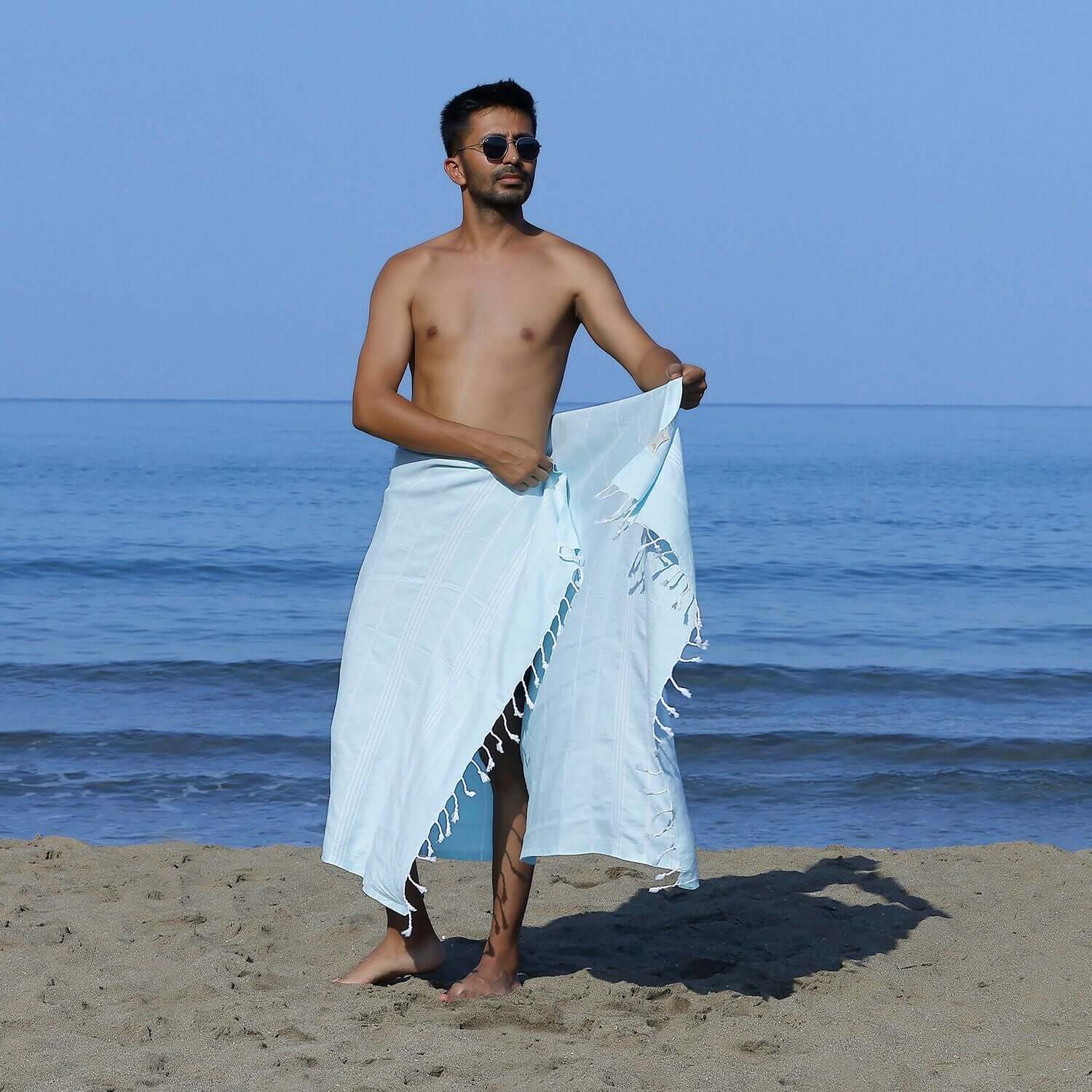 Man at the beach showcasing a Loom Legacy aqua striped beach towel with tassels, with the sea in the background.