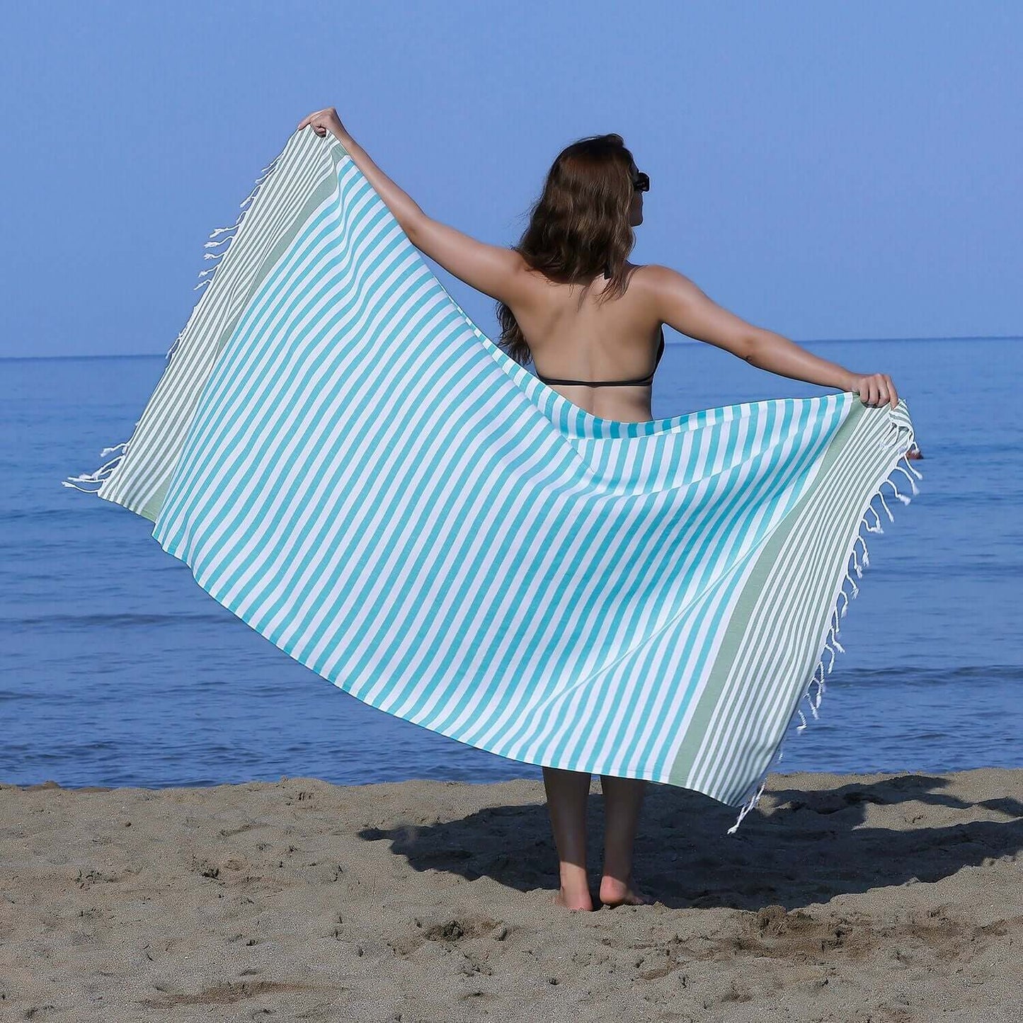  Woman at the beach showcasing a Loom Legacy turquoise and green striped beach towel with tassels, with the sea in the background.