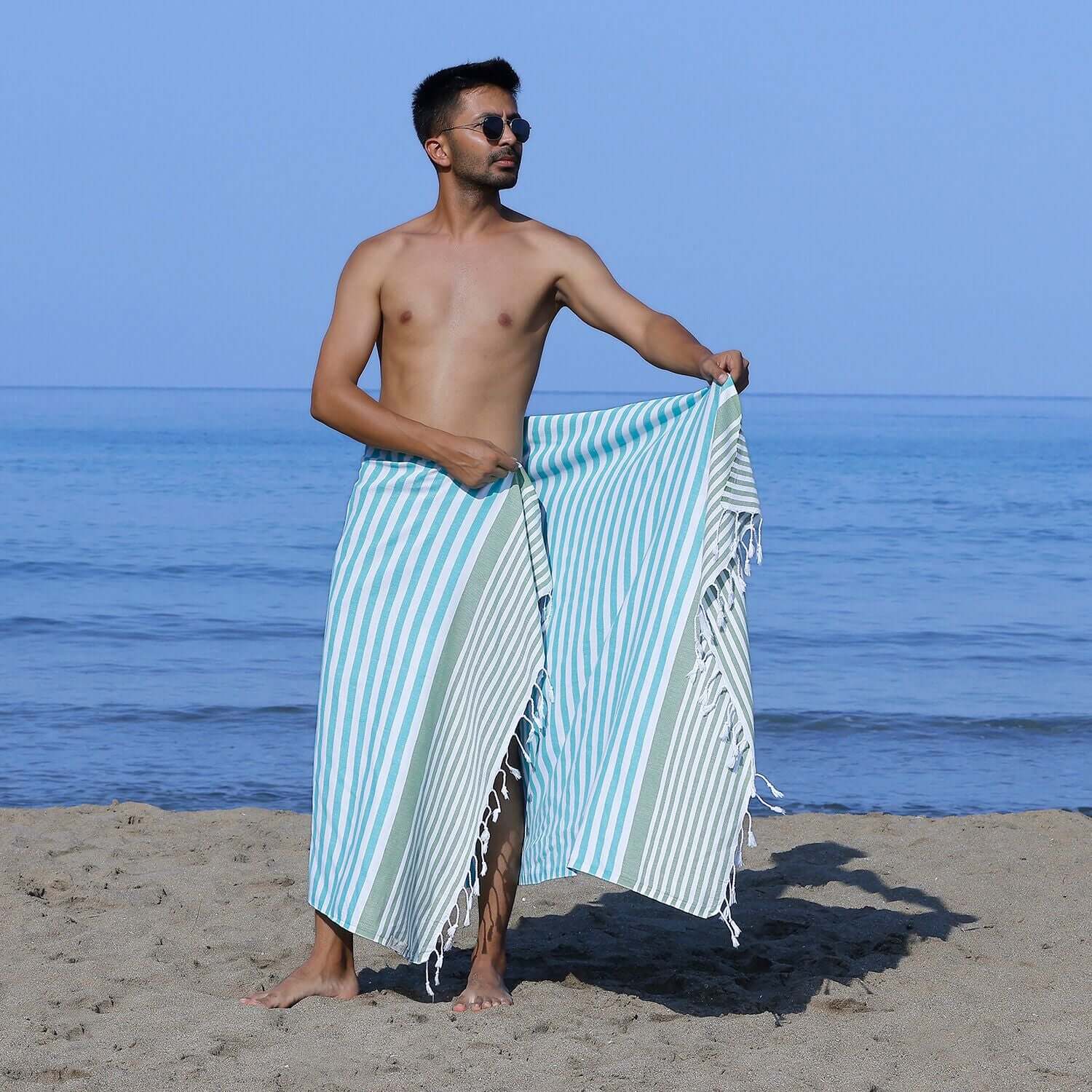 Man at the beach showcasing a Loom Legacy turquoise and green striped beach towel with tassels, with the sea in the background.