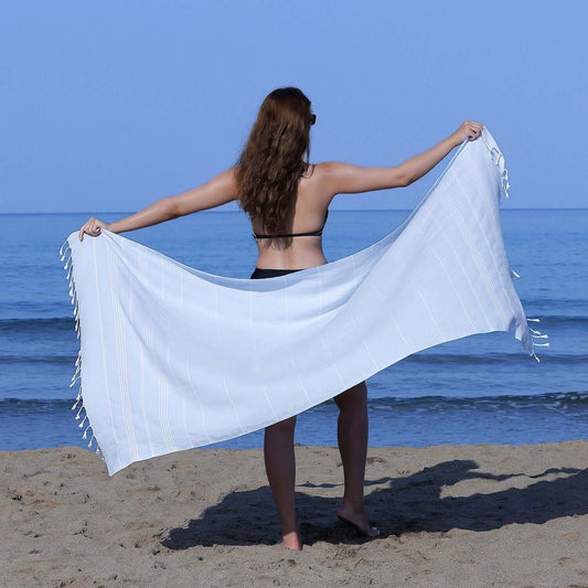 Woman at the beach showcasing a Loom Legacy light blue striped beach towel with tassels, with the sea in the background.