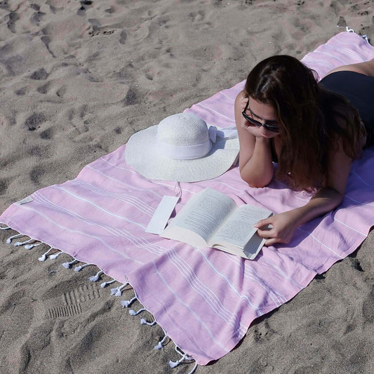 Woman relaxing on a sandy beach, lying on a Loom Legacy pink and white striped beach towel with tassel details, paired with a white sun hat and reading a book.