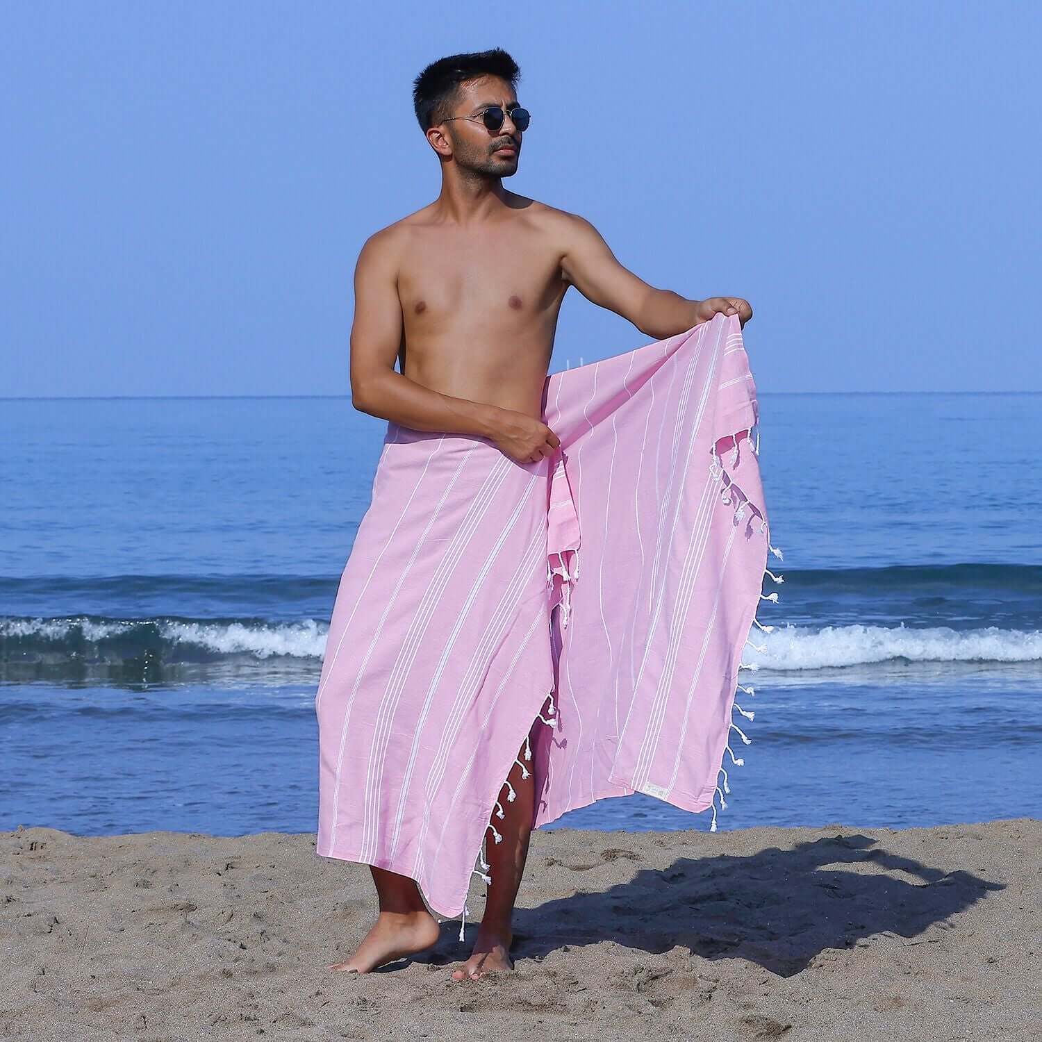 Man at the beach showcasing a Loom Legacy pink striped beach towel with tassels, with the sea in the background.