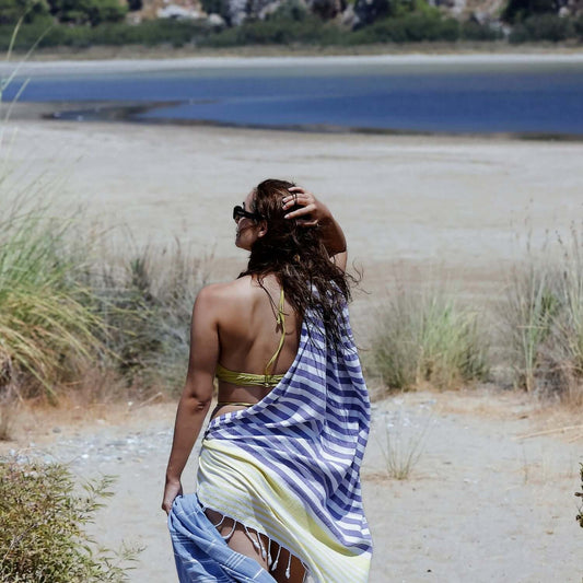 Woman gazing at a serene lakeside view, draped in a Loom Legacy purple and yellow striped beach towel with tassel accents, complemented by her green bikini and stylish sunglasses.