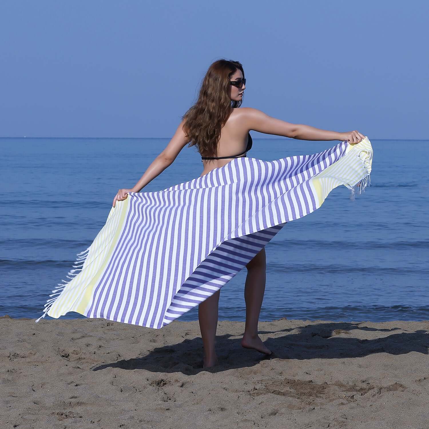 Woman at the beach showcasing a Loom Legacy purple and yellow striped beach towel with tassels, with the sea in the background.