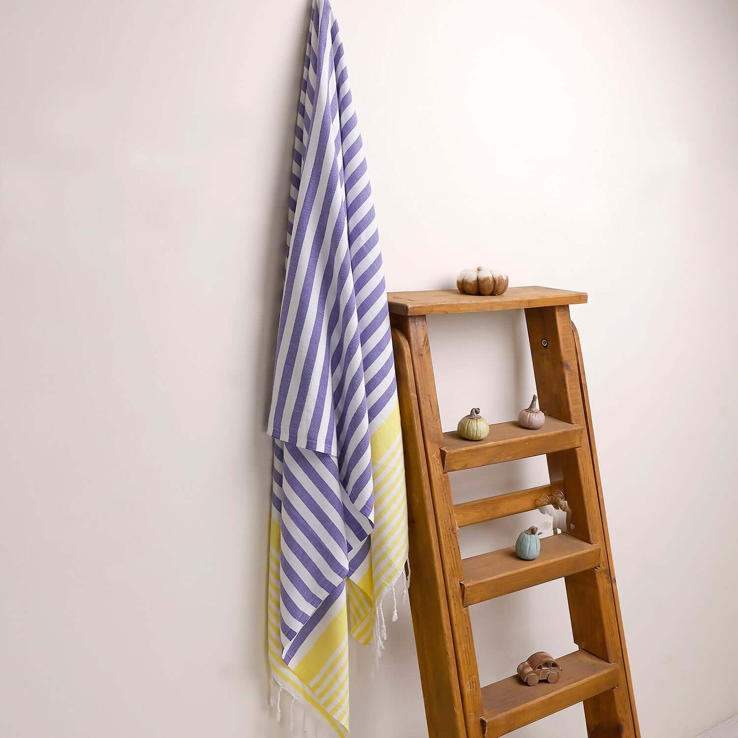 Loom Legacy purple and yellow striped beach towel with tassels, draped over a wooden ladder beside small decorative items on the shelves.